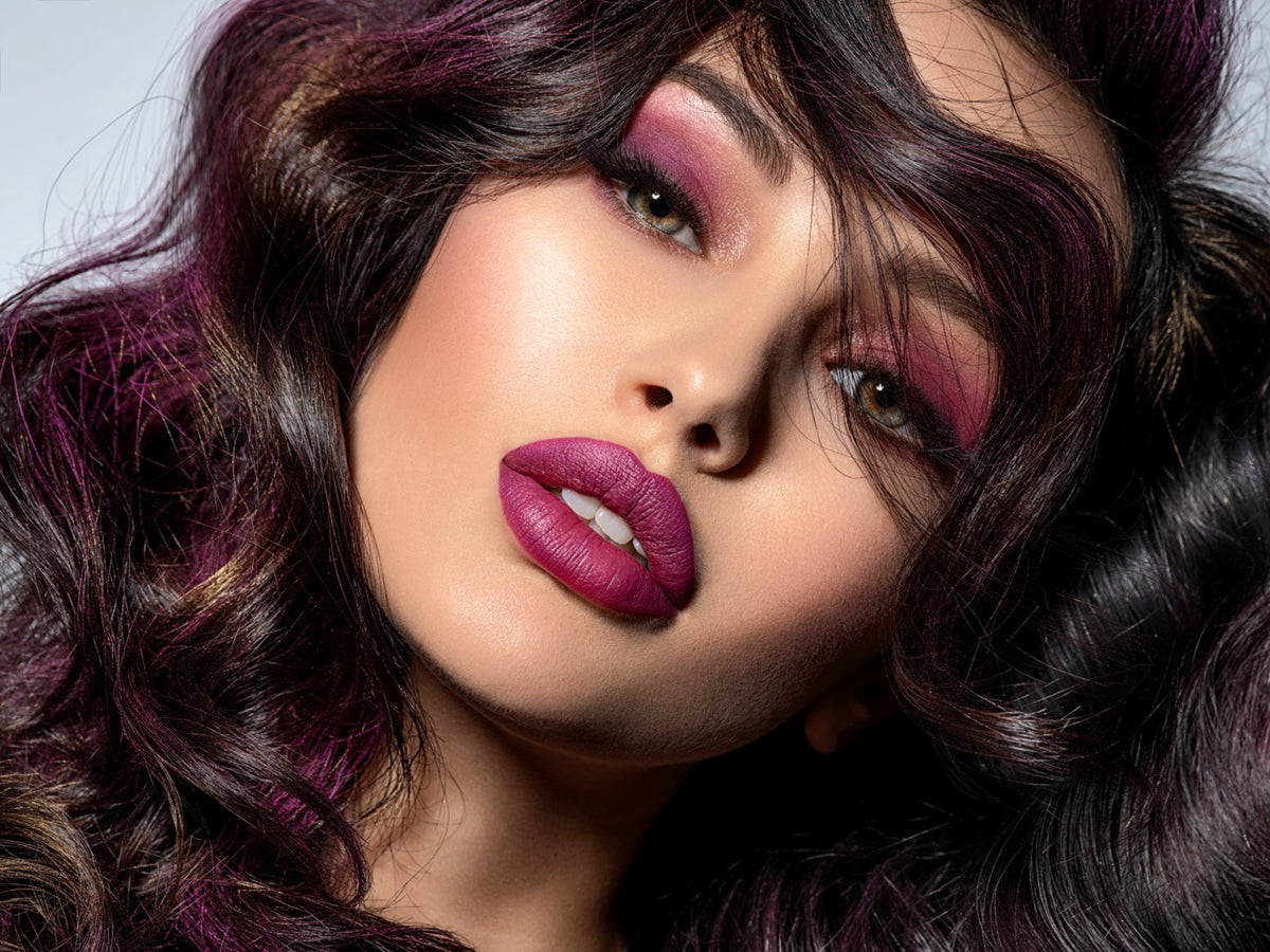 Close-up portrait of a woman with voluminous dark wavy hair featuring prominent purple and magenta highlights, styled with matching purple eyeshadow and lipstick against a light gray background.