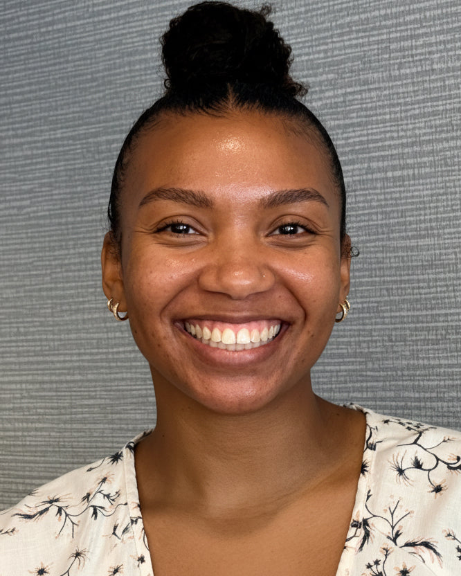 A smiling woman with her hair in a high bun wearing a white floral top, standing against a gray textured wall.
