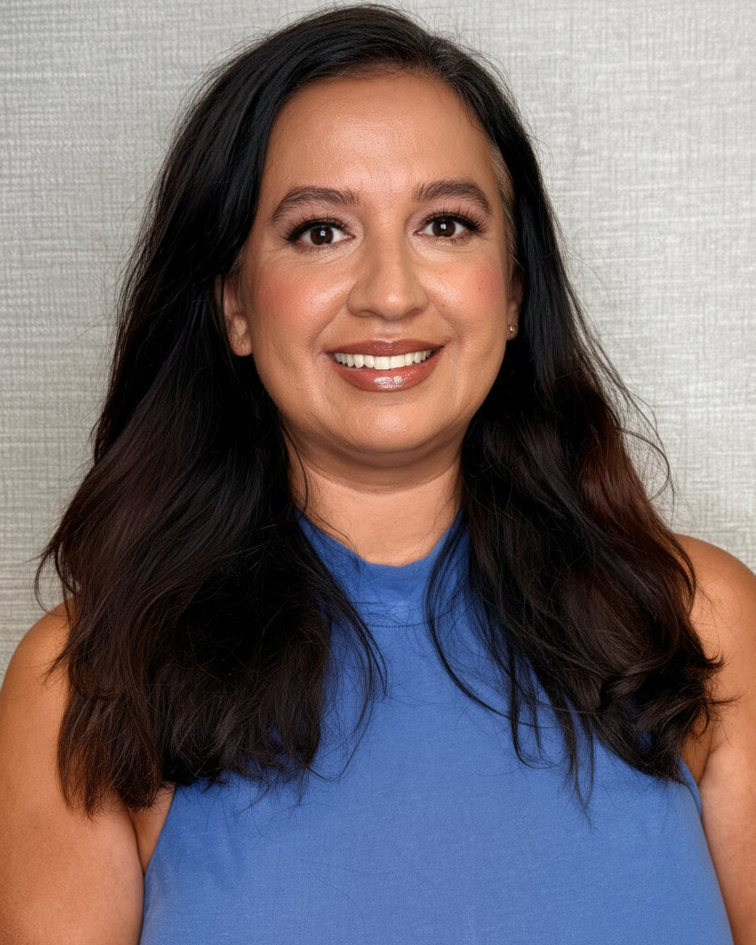 A smiling woman with long dark hair wearing a sleeveless blue top, standing against a textured light gray wall.