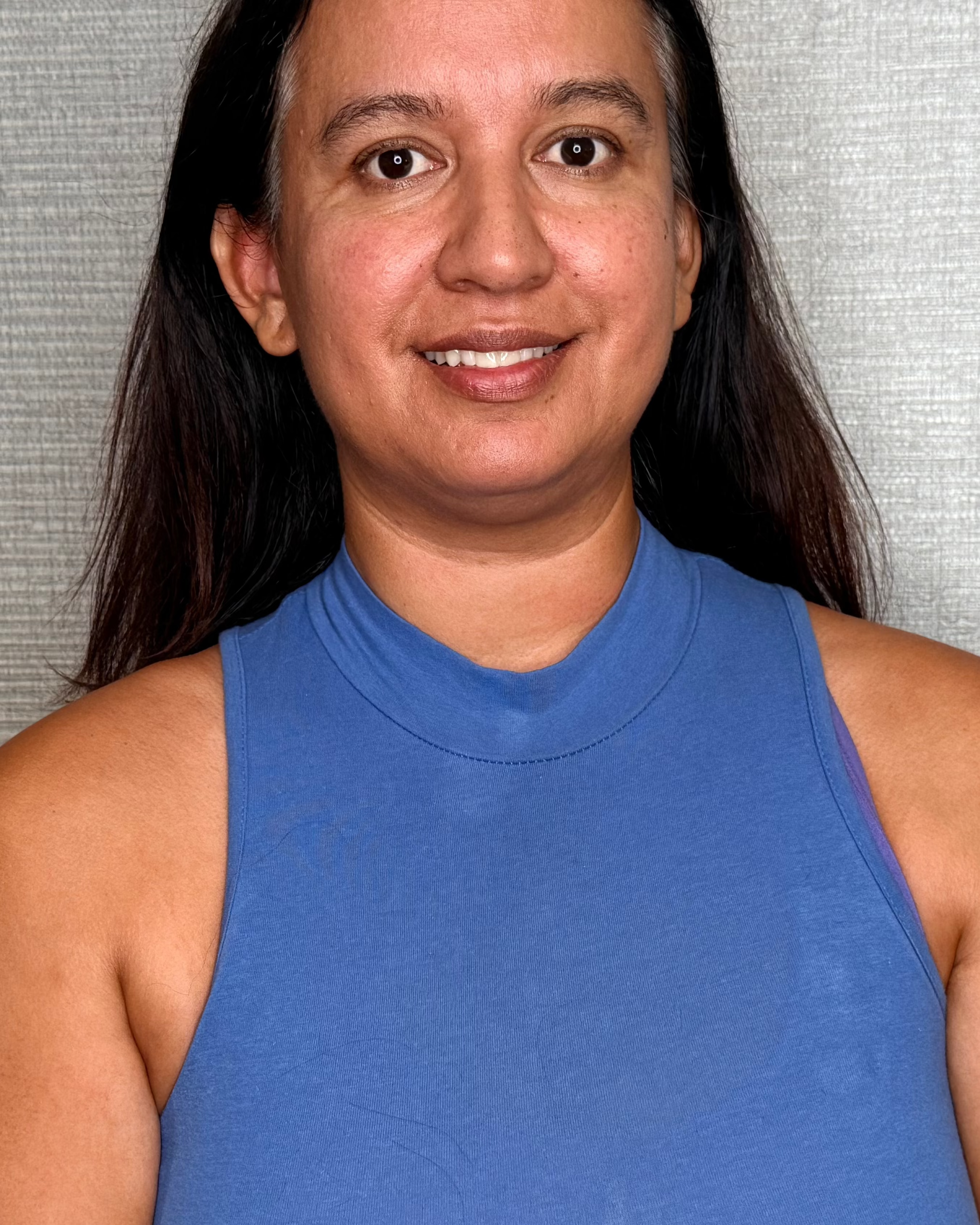 A woman with long dark hair wearing a blue ribbed sleeveless mock-neck top, standing against a textured gray wall.