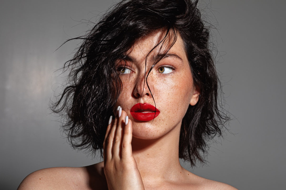 A close-up portrait of a woman with messy dark hair, freckles, and bright red lipstick against a plain gray studio background. No product packaging or brand logo is visible.
