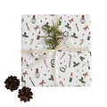 A gift box wrapped in white Christmas-themed paper with candy canes, holly, stars, and lipstick illustrations, tied with a cream ribbon and topped with a green pine sprig. Two pinecones sit on the light beige surface beside the box.