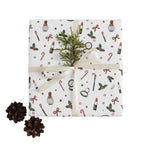 A gift box wrapped in white Christmas-themed paper with candy canes, holly, stars, and lipstick illustrations, tied with a cream ribbon and topped with a green pine sprig. Two pinecones sit on the light beige surface beside the box.