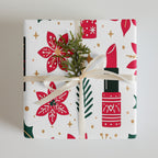 A square gift box wrapped in white holiday-themed paper with red poinsettias, gold stars, green leaves, and a red lipstick illustration, tied with a cream ribbon and topped with a small green sprig, on a light gray background.